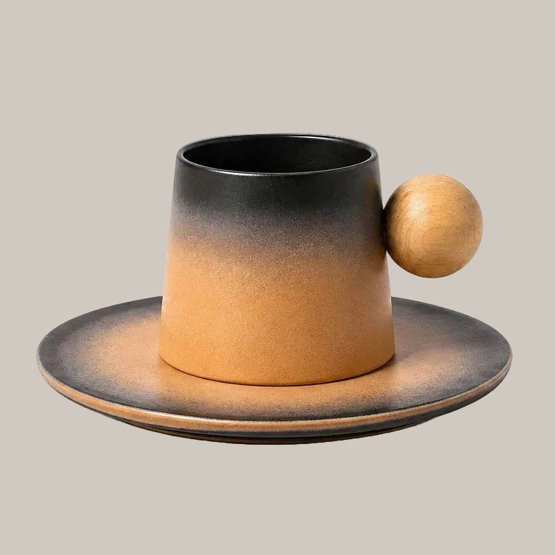 A ceramic cup and saucer set with a matte black-to-tan gradient glaze and a small wooden ball accent on the side, shown on a plain warm gray background.