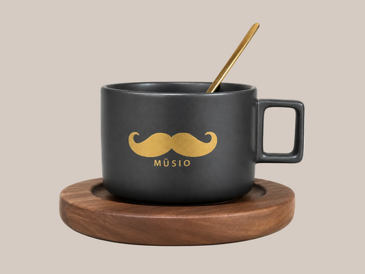 A matte black ceramic mug with a gold mustache graphic and the word 'MÜSIO' printed below, shown on a round wooden coaster with a gold spoon resting inside, against a plain white background.